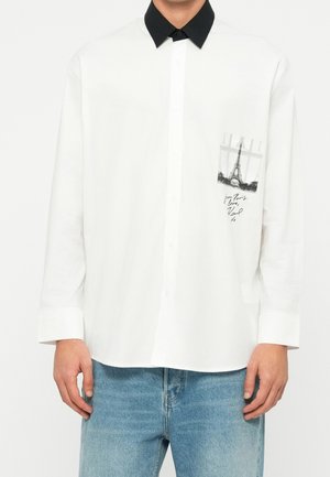 Hvid langærmet skjorte med sort krave, med et lommeprint af Eiffeltårnet og håndskrevet tekst, båret med blå jeans.