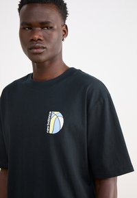 Black cotton t-shirt with short sleeves, featuring a colorful logo design on the left chest: blue, yellow, and white accents.