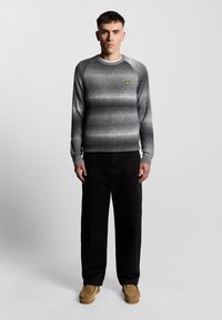 Grauer Strickpullover mit einem gerippten Muster, Rundhalsausschnitt und einem gelben Emblem auf der Brust, kombiniert mit schwarzen Hosen und braunen Schuhen.
