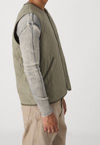 Man som bär en olivgrön vadderad väst över en grå ribbad långärmad skjorta och beige byxor, visad från sidan mot en vit bakgrund.