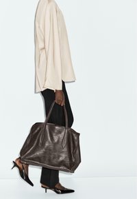 Sac en cuir marron avec deux poignées, texture lisse, forme rectangulaire, accompagné d'un haut oversize beige clair et d'un pantalon noir.
