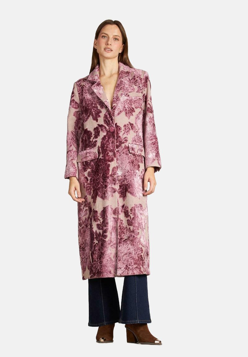 Long manteau rose à motif floral avec col, poches avant et coupe droite, porté sur un pantalon évasé bleu et des talons marron.