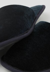 Schwarze Cordruy-Schlappen mit einer strukturierten Oberfläche, runder Form und kontrastierendem dunklem Stoffbesatz. Weiches Material und minimale Nahtakzente.