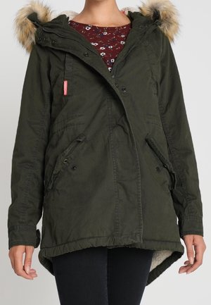Veste parka vert foncé avec capuche bordée de fourrure, poches avant et fermetures à boutons-pression, portée sur un top rouge à fleurs.