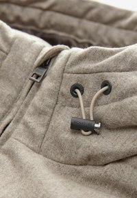 Beige strukturierter Jackenstoff mit Reißverschluss, ausgestattet mit schwarzen Ösen und einem beigen Zugband mit einem markenprägten Stopper.