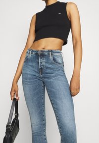 Top corto negro con textura acanalada, combinado con jeans de mezclilla rectos de color azul claro. La modelo lleva un bolso negro con una correa desmontable.
