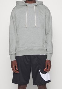 Personne portant un sweat à capuche gris clair avec cordons et un short de sport noir avec un grand logo blanc Nike Swoosh sur la jambe gauche.
