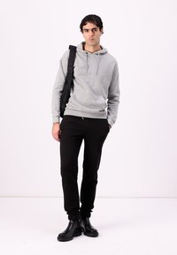 Un sweat à capuche gris avec une poche kangourou, associé à un pantalon noir ajusté et des bottes noires volumineuses. Le modèle tient un sac noir, devant un fond uni.