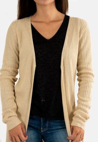 Una donna indossa un cardigan a coste beige sopra una maglietta nera a V e jeans blu, con le mani che tengono i bordi del cardigan.
