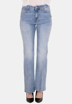 Donna che indossa jeans azzurri a gamba dritta e tacchi neri con punta a punta, in piedi davanti a uno sfondo semplice.