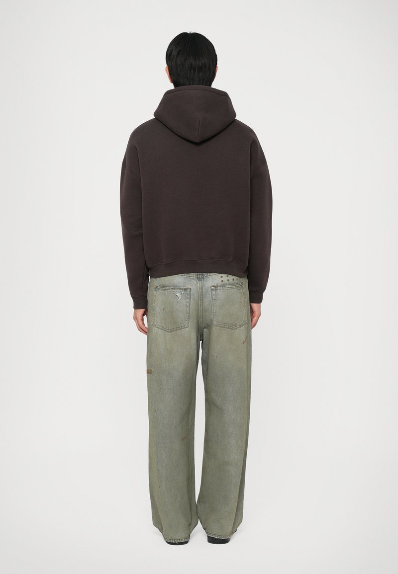 Felpa cappuccio marrone scuro con un design cropped, abbinata a jeans a gamba larga verde chiaro con dettagli strappati e cuciture visibili.