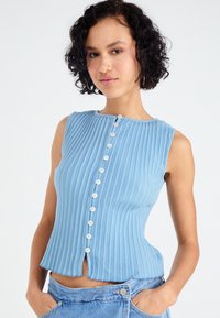 Lichtblauwe mouwloze top met verticale ribbelstructuur, met een hoge halslijn en centrale knoopsluiting, iets uitlopend aan de onderkant.