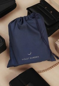 Navy-taustalla oleva narupussi, jossa on kultainen "VIOLET HAMDEN" -teksti ja puolikuun yksityiskohta, ympäröity mustilla nahkalaukuilla.