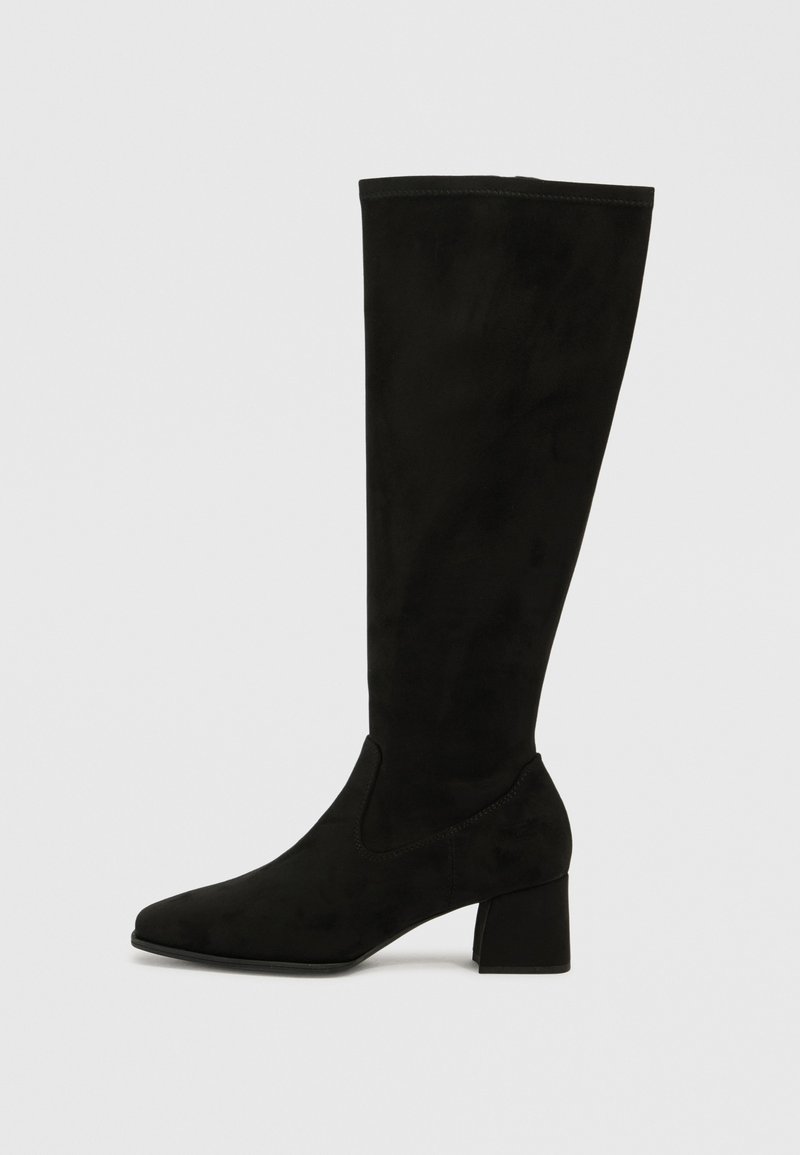 Bota de ante negra hasta la rodilla con puntera afilada y tacón bloque. Presenta una textura suave y paneles laterales elásticos para facilitar su uso.