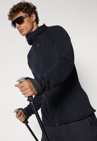 Schwarze zip-up Jacke mit strukturiertem Finish, hochstehendem Kragen, Daumenlöchern und sportlichem Schnitt. Hält Trekkingstöcke mit grauen Griffen.
