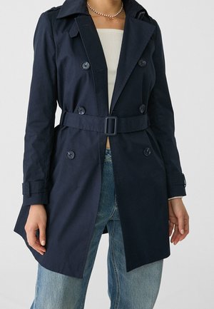 Kobieta ubrana w granatowy płaszcz trencz z paskiem, na białym prążkowanym topie i jasnoniebieskich dżinsach, z różowym naszyjnikiem z pereł.