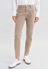 Pantalones de pana beige con un corte ajustado, que cuentan con bolsillos frontales, pasadores para cinturón y cierre de botón. Combinados con zapatillas blancas.