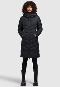 Doudoune noire avec capuche, présentant un motif à chevrons, fermeture éclair complète et cordons réglables. Associée à des bottes de combat noires.