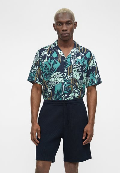 Άνδρας με κοντά ξανθά μαλλιά που φορά μπλε, πράσινο και καφέ πουκάμισο με τροπικό μοτίβο, κοντό μανίκι και μαύρα υφασμάτινα σορτς, στέκεται μπροστά σε λευκό φόντο.