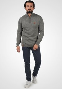 Grijze gebreide trui met een hoge kraag en een halve rits, voorzien van geribbelde accenten bij de zoom en manchetten, gecombineerd met marineblauwe broek en witte sneakers.