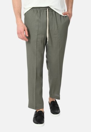 Uomo con pantaloni verde oliva con coulisse e scarpe slip-on nere con suola bianca, in piedi con una mano in tasca.