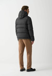 Jachetă tip puffer de culoare neagră, cu glugă, având un design matlasat și manșete elastice. Purtați peste pantaloni bej și încălțări negre.