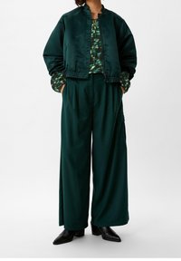 Grüne Satin-Bomberjacke mit Stehkragen, kombiniert mit weit geschnittenen grünen Hosen mit Seitentaschen, getragen mit schwarzen Heels.