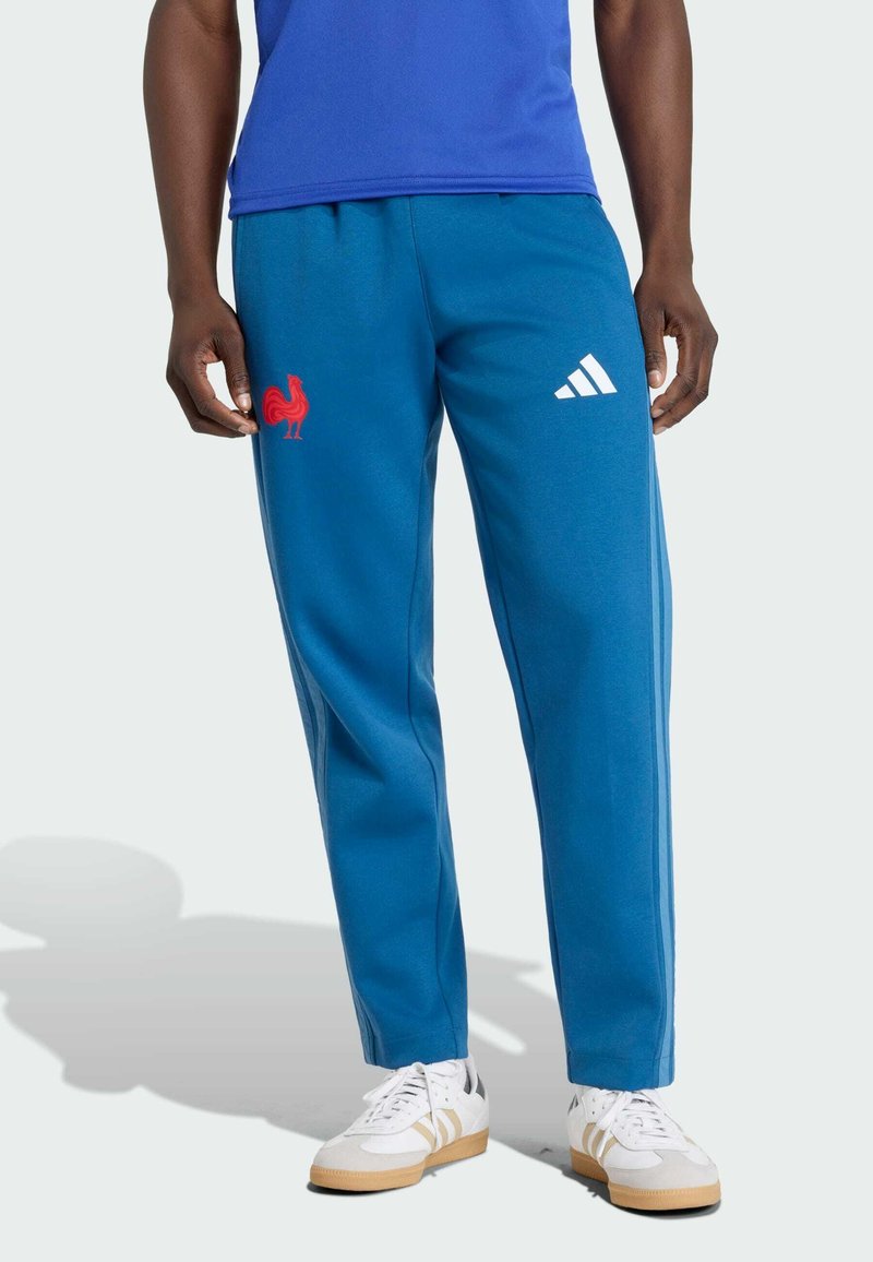 Modré tepláky s červeným logom kohúta na ľavom stehne a bielymi prúžkami Adidas na pravej strane, vyrobené z hladkého materiálu, voľný strih.