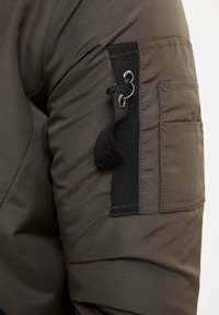 Giacca verde oliva con tessuto testurizzato, tasca con aletta sulla spalla e cinturino in nylon nero con anello metallico per la chiusura.