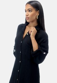 Abito camicia nero, con texture, maniche lunghe, chiusura a bottoni e una tasca. Il tessuto sembra morbido e leggero, con polsini arrotolati.