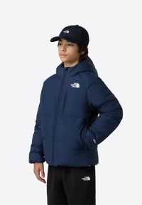 Tumesinine taskurätiga sulejope tõmblukuga ja kapuutsiga, rinnal valge logo ja elastsed varrukapitsid. Kandmisel mustad püksid.