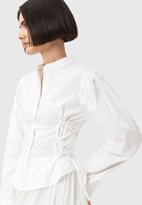 Camicia di cotone bianca con colletto alla mandarino, maniche a sbuffo e dettagli in pizzo in vita. Tessuto liscio e orlo curvato.