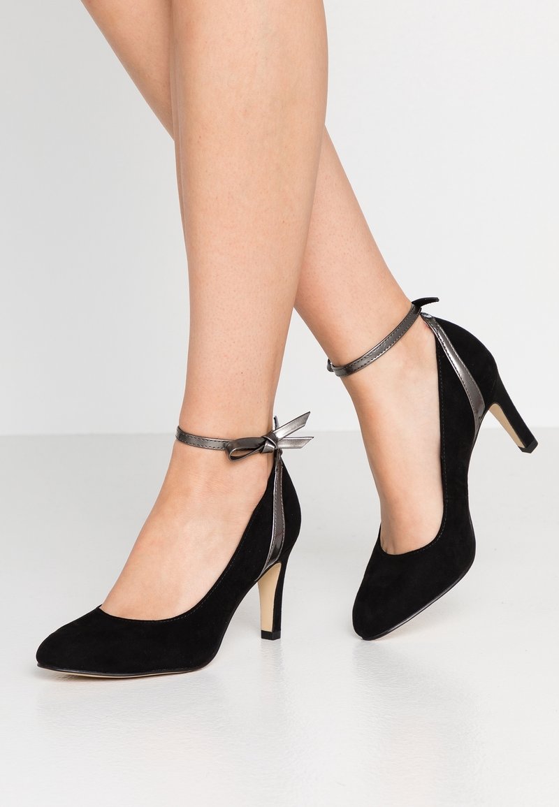 Chaussures à talons hauts en daim noir avec un bout rond, une bride métallique à la cheville et un détail nœud. Elles présentent un talon fin et une texture lisse.