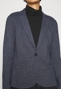 Námořnická modrá blazer z texturovaného materiálu, s jedním knoflíkovým zapínáním, klopovým límcem a dvěma předními kapsami. Nositá přes černý rolák.
