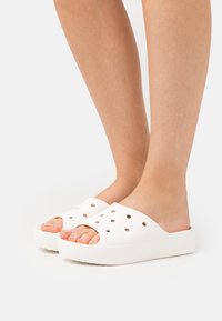 Mules blancs à enfiler fabriqués à partir de matériaux durables, présentant un design à bout ouvert, des trous de ventilation et une semelle épaisse.