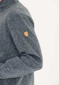 Grå texturerad jacka med hög krage och ett emblem på vänster axel. Har kontrasterande sömmar och mjukt, stretchigt tyg.