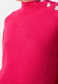 Gros plan sur un pull à col roulé côtelé rose vif avec trois ornements décoratifs en forme de cœur argenté sur l'épaule.