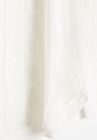 White blouse made of lightweight fabric, featuring a button placket and a rounded hem. The texture is smooth with a subtle sheen.