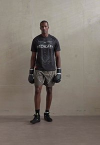 Joven vestido con guantes de boxeo negros, camiseta gráfica negra y pantalones cortos oliva, de pie contra una pared de concreto liso.
