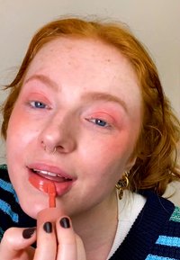 Ferskenfarget leppeglansapplikator i hånden, påfører den på lyse rosa lepper. Har lett fregnet hud, blå øyne og rødt hår med en stripete genser.