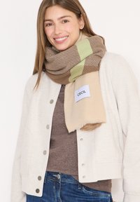 Beige og brunt stribet halstørklæde med grønne accenter, båret over en lysegrå jakke og en brun sweater, der viser bløde teksturer og en etiket.