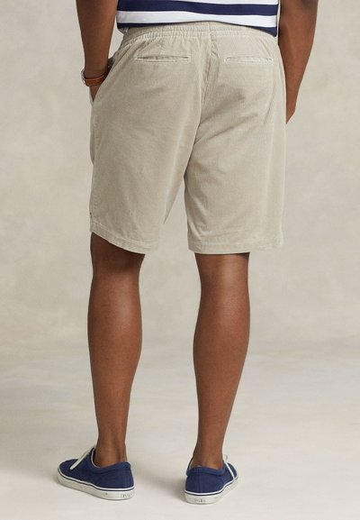 Μπεζ βερμούδα από κορντόρι με ελαστική μέση και πίσω τσέπες, εμφανιζόμενη από πίσω. Φοριέται με μπλε καμβά παπούτσια.