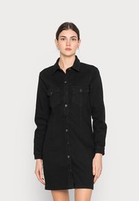 Vestido de camisa em denim preto com fecho frontal de botões, dois bolsos no peito, mangas longas e uma silhueta justa com comprimento até à metade da coxa.