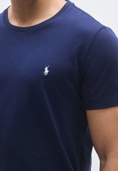 Tmavomodré bavlnené tričko s okrúhlym výstrihom a krátkymi rukávmi. Na ľavej strane hrudníka je malý biely vyšívaný logo. Hladká textúra.
