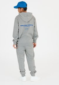 Grijze sweatshirt en bijpassende joggers gemaakt van zacht materiaal. De sweatshirt heeft op de rug blauwe tekst "NEW BAD HABITS" en het logo "LES'ARCS".