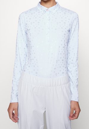 Lyseblå langærmet poloshirt med små stjernemønstre, båret med løstsiddende hvide bukser, vist fra nakke til hofter, hænder afslappet ned langs siderne.