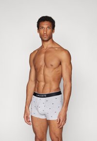 Gray boxer shorts with a soft texture and small black patterned accents. Features a black waistband with "LACOSTE" logo. Model standing, facing forward.