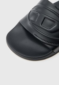 Czarna klapka na płaskiej podeszwie z teksturowanym paskiem, charakteryzująca się szerokim, formowanym fasonem i gładką, zaokrągloną podeszwą. Materiał wydaje się syntetyczny.