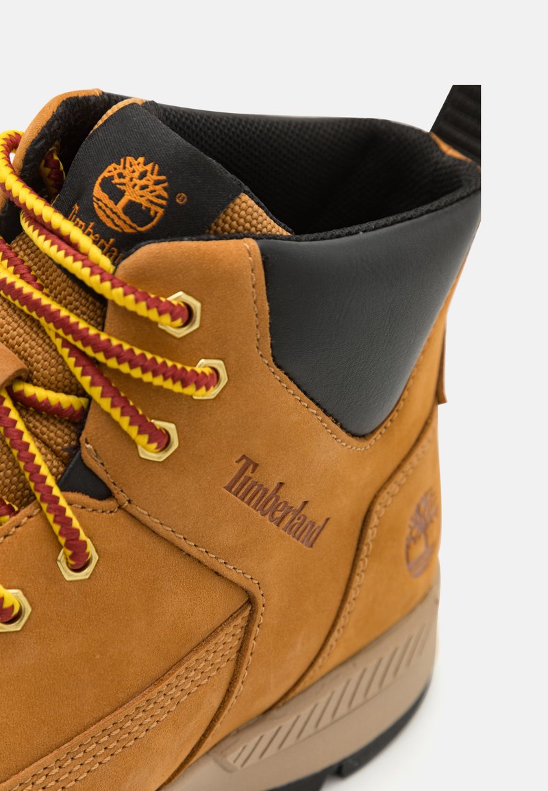 Bota Timberland con parte superior de ante color marrón claro, acentos de cuero negro, cordones amarillos y rojos, ojales dorados y suela de goma texturizada.