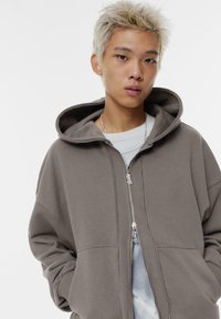 Felpa con zip di grandi dimensioni, marrone, dal tocco morbido, con un ampio cappuccio e tasche frontali. Indossata sopra una camicia a righe blu chiaro.